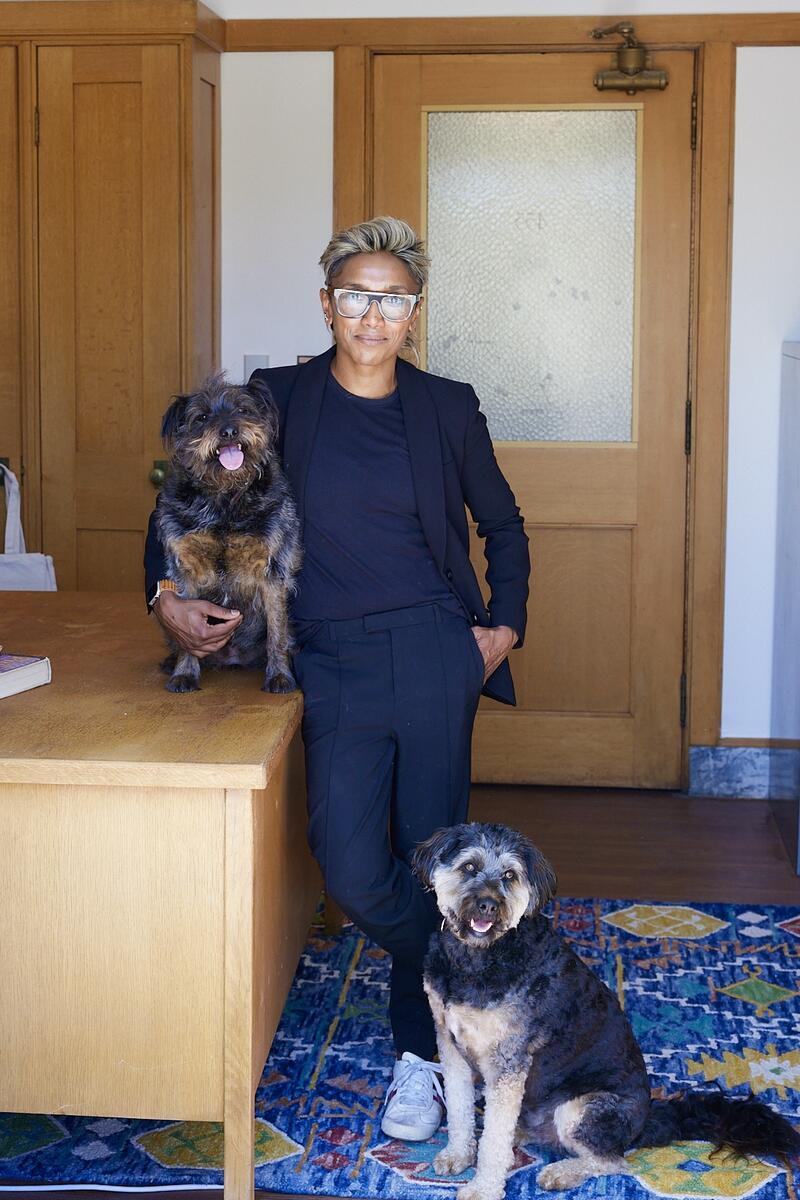 Poulomi Saha with their two dogs. Saha is standing upright indoors and smiling. They are wearing a dark suit and matching dark collarless shirt. They are a scholar of Asian American literature, postcolonial studies, and queer and feminist theory.